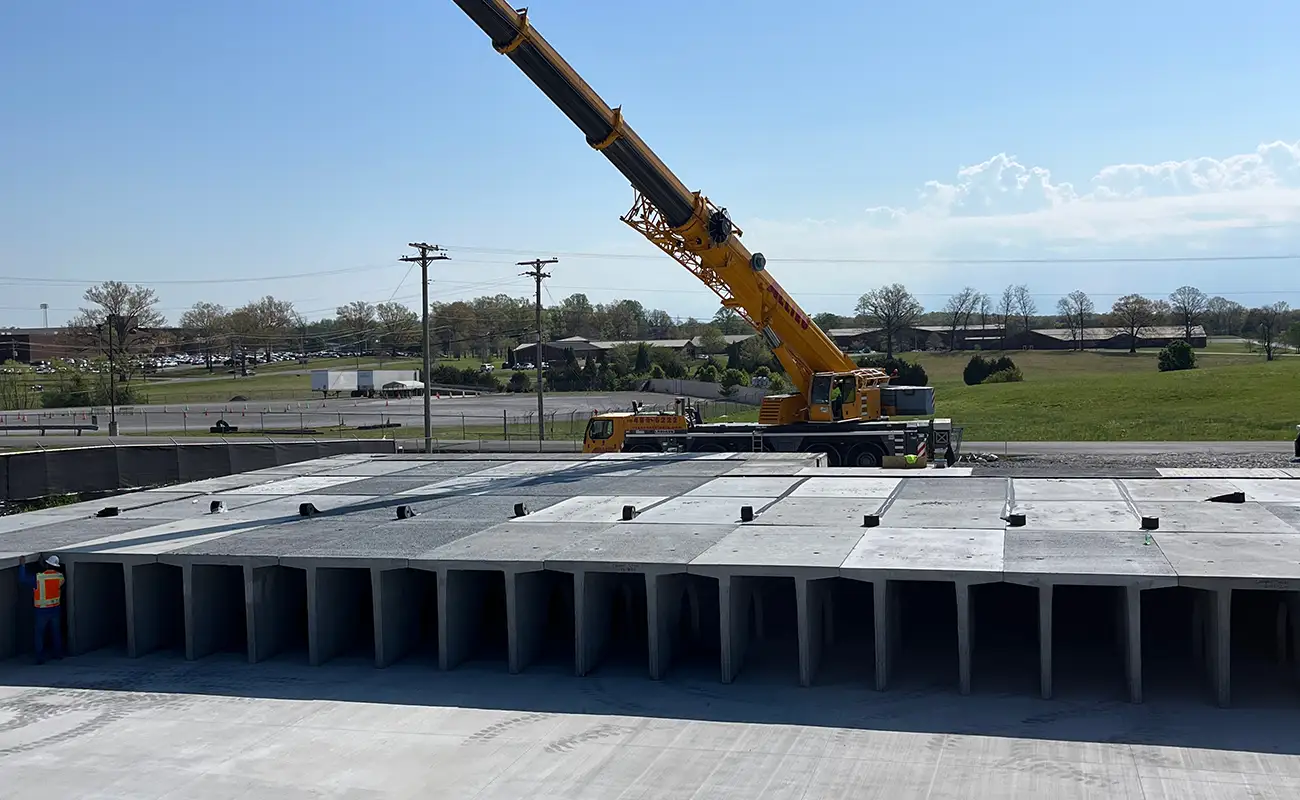 A large yellow crane lifts concrete panels onto a grid of rectangular concrete structures at an outdoor construction site. Two workers in safety vests stand nearby.