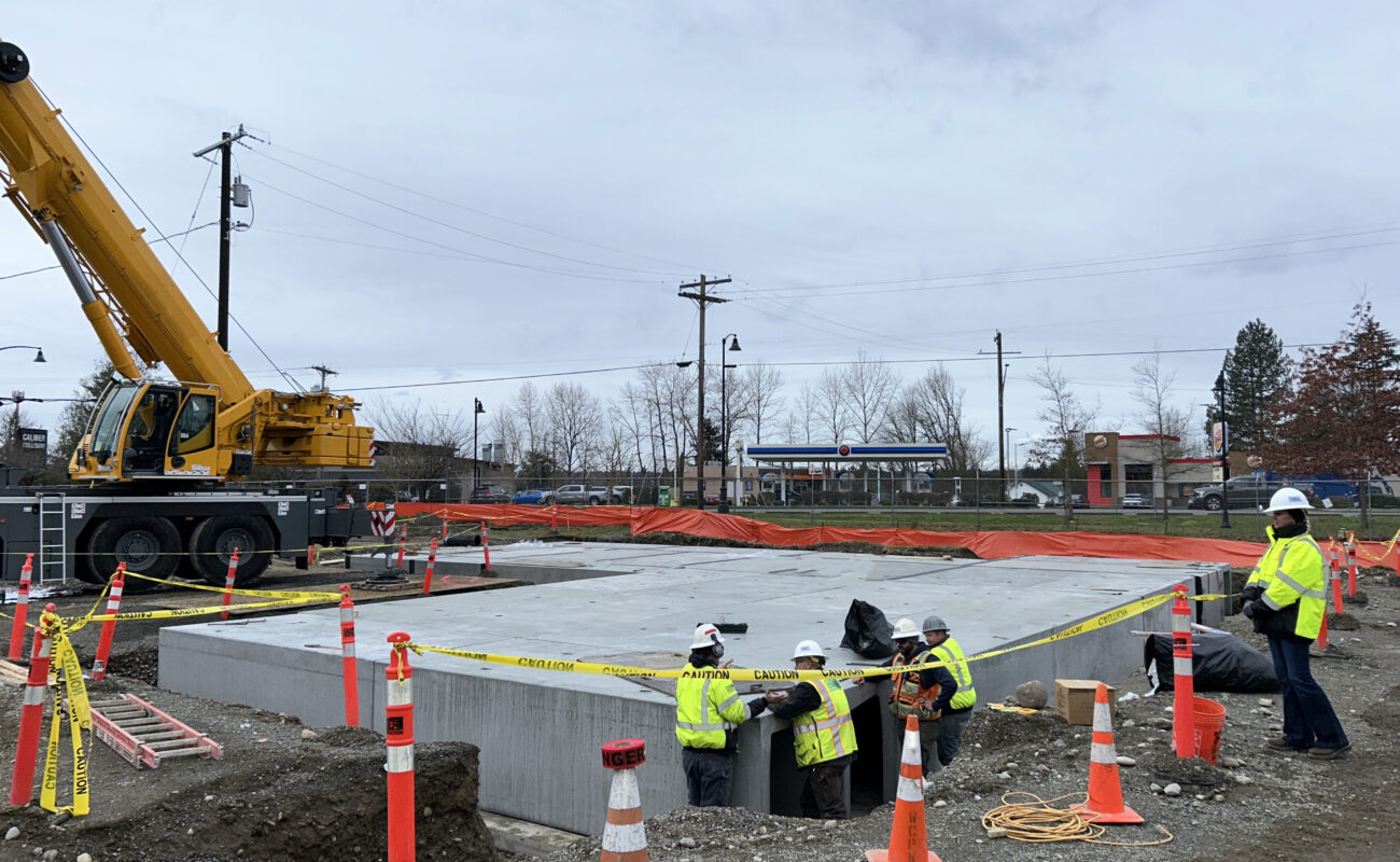 Construction workers in safety gear work around a large concrete foundation at a site with a crane, caution tape, and traffic cones on an overcast day.