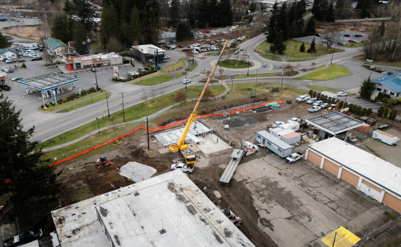 A construction site with a yellow crane lifting materials near a road, surrounded by buildings, vehicles, and trees on a cloudy day.