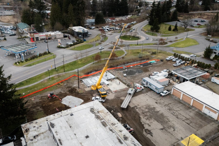 A construction site with a yellow crane lifting materials near a road, surrounded by buildings, vehicles, and trees on a cloudy day.