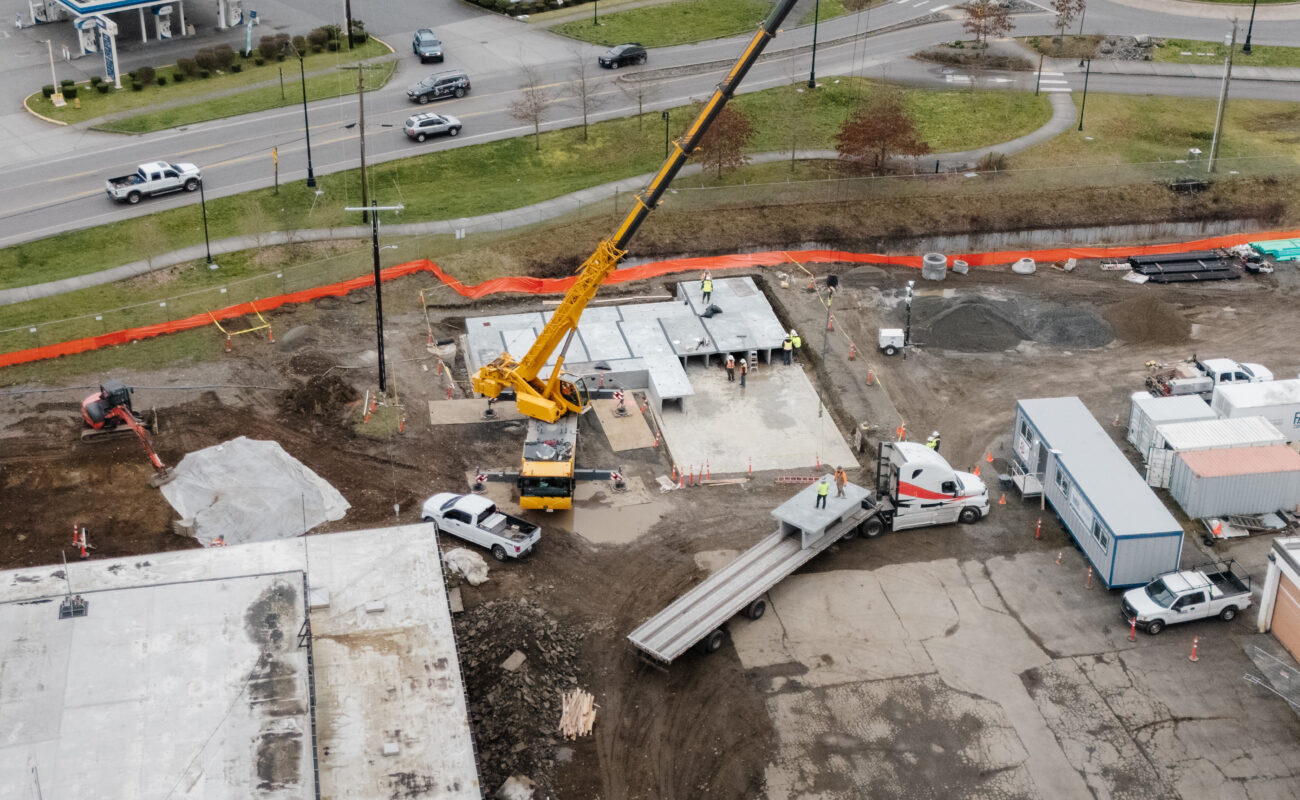 Aerial view of a construction site with a crane lifting materials, trucks, and workers present; nearby road and parked vehicles visible.