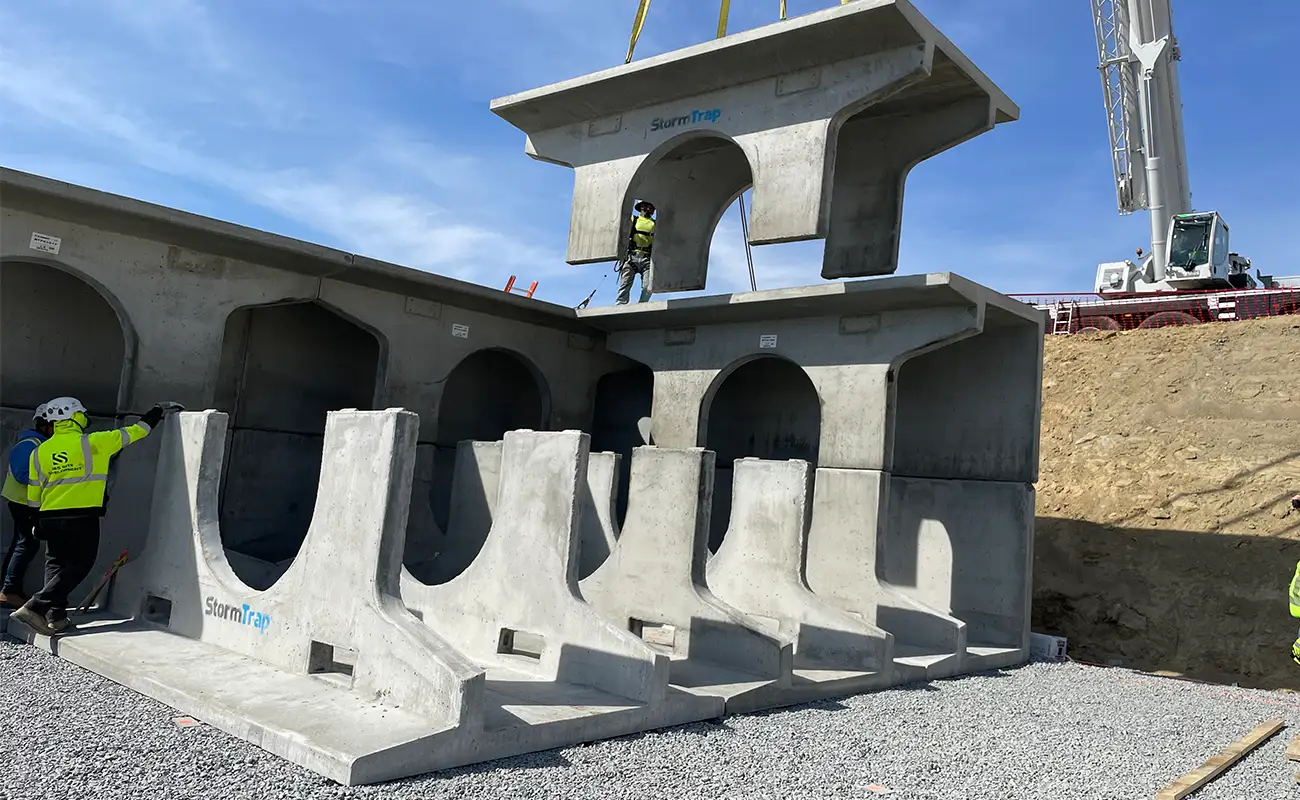 Workers assemble large precast concrete sections with cranes at a construction site, stacking tunnel-like components on a gravel surface.