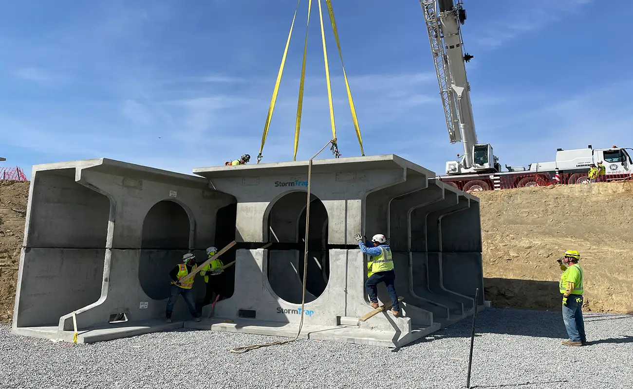 Construction workers guide large concrete structures being lifted by a crane at a construction site with gravel ground and earth-moving equipment.
