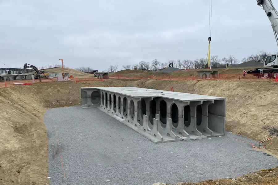 A large concrete culvert section is placed on a gravel bed in an excavated area at a construction site, with a crane and workers visible in the background.