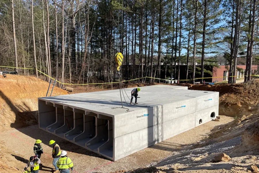 Construction workers guide the placement of a large precast concrete stormwater structure in a wooded area, with four visible entryways and safety equipment in use.
