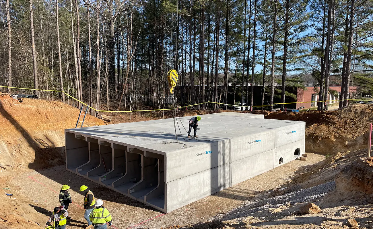 Construction workers guide the placement of a large precast concrete stormwater structure in a wooded area, with four visible entryways and safety equipment in use.