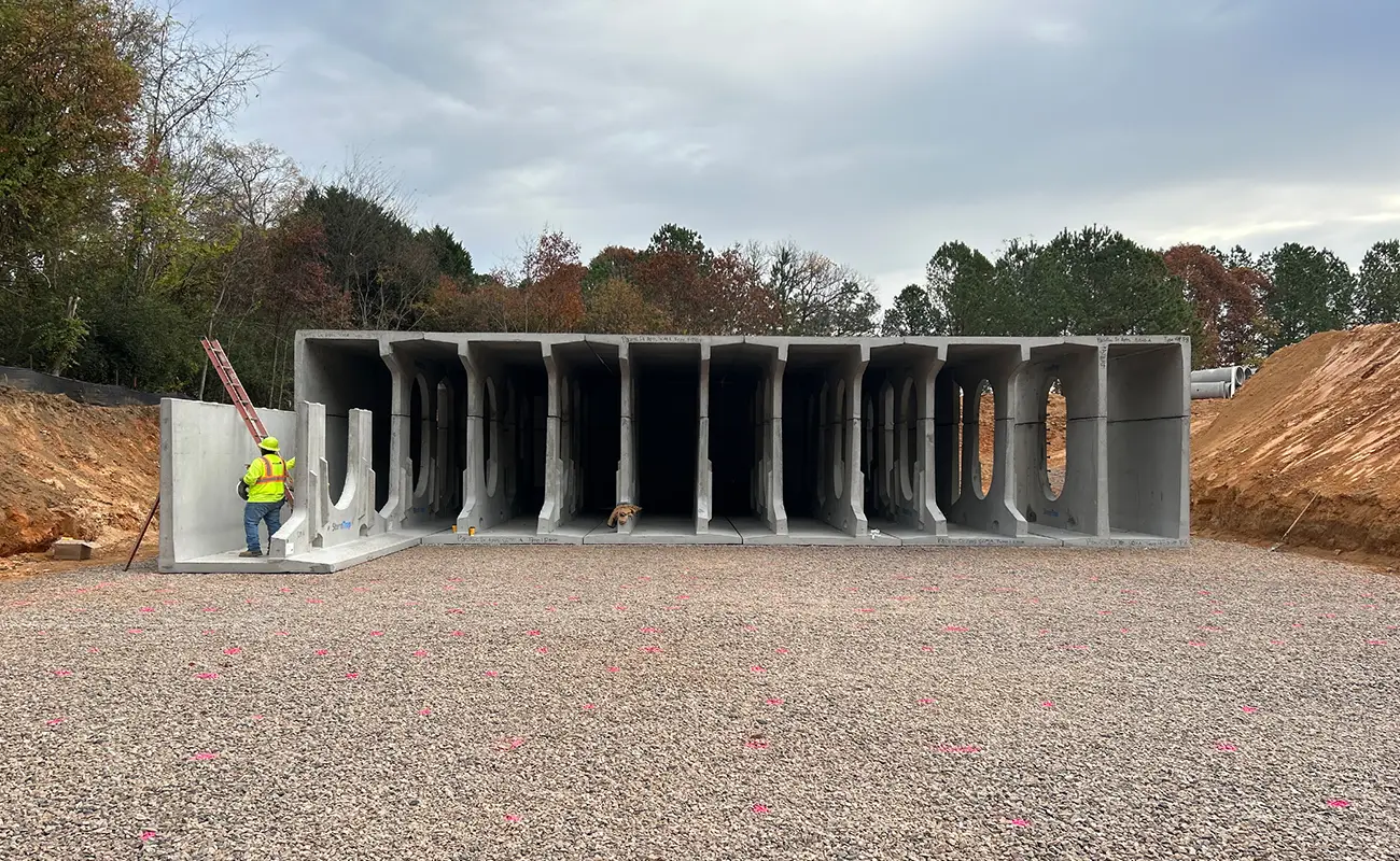 A construction worker stands beside large concrete box culverts arranged in rows at a worksite, with dirt, gravel, and trees in the background.