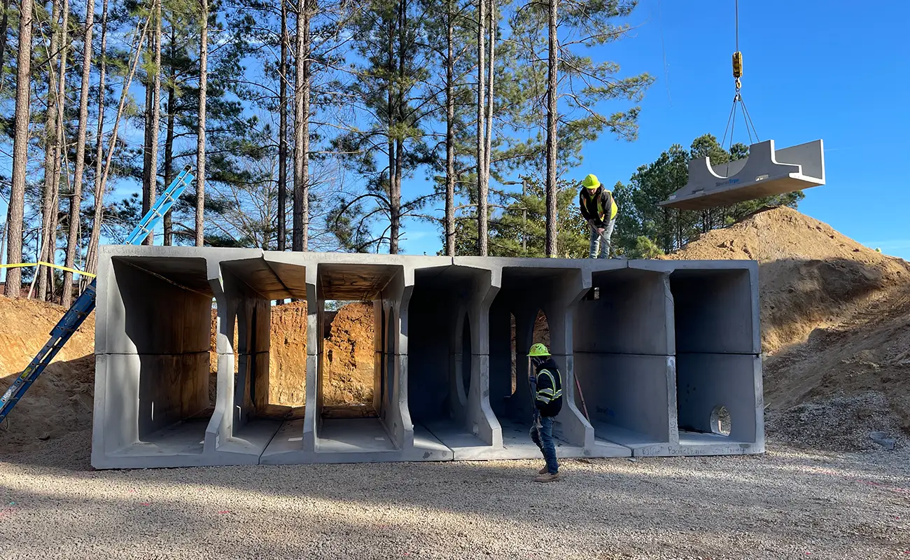 Three construction workers assemble large concrete box culverts at a work site with trees and construction equipment in the background.