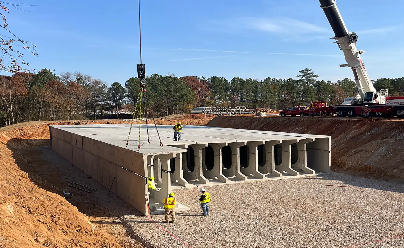 Construction workers guide a large precast concrete culvert section being lowered by a crane onto a gravel foundation at a construction site surrounded by trees.