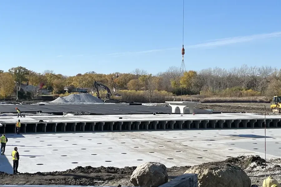 Construction workers operate machinery and install large concrete panels at a building site under clear blue skies, with trees and equipment visible in the background.