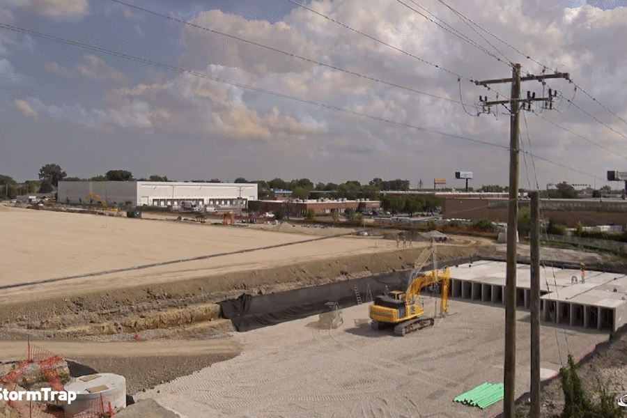 A construction site with an excavator working near large drainage pipes, surrounded by dirt, gravel, and some buildings in the background under a partly cloudy sky.