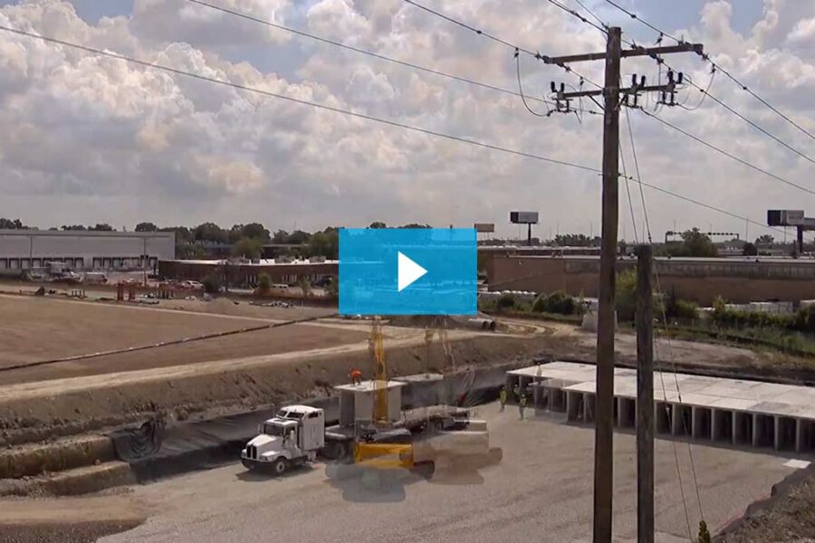 Construction site with trucks and equipment, power lines in foreground, storage units or concrete structures on the right, and industrial buildings in the background under a partly cloudy sky.