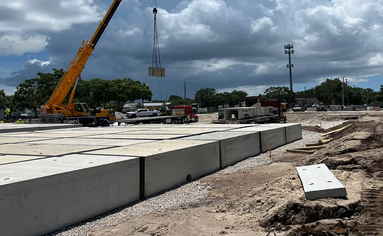 A construction site with a crane lifting a large concrete slab, several trucks, and workers present under a partly cloudy sky.