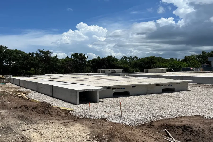 A construction site with large concrete slabs placed on a gravel foundation, surrounded by dirt and trees under a partly cloudy sky.