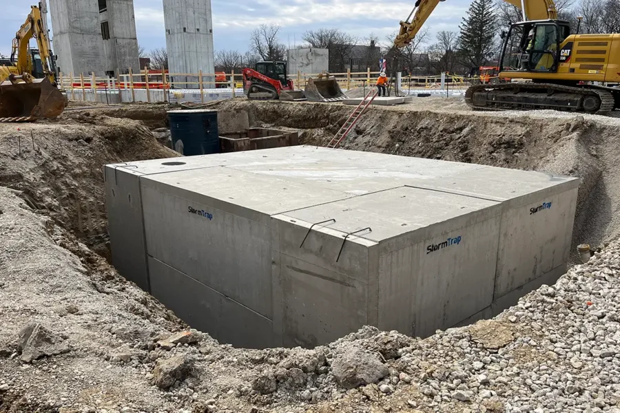 A large precast concrete vault is partially buried in a construction site with heavy machinery and building materials nearby.