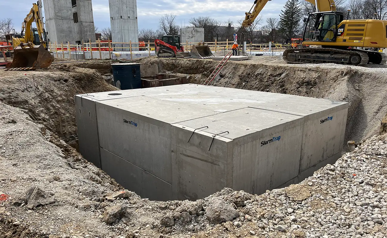 A large precast concrete vault is partially buried in a construction site with heavy machinery and building materials nearby.
