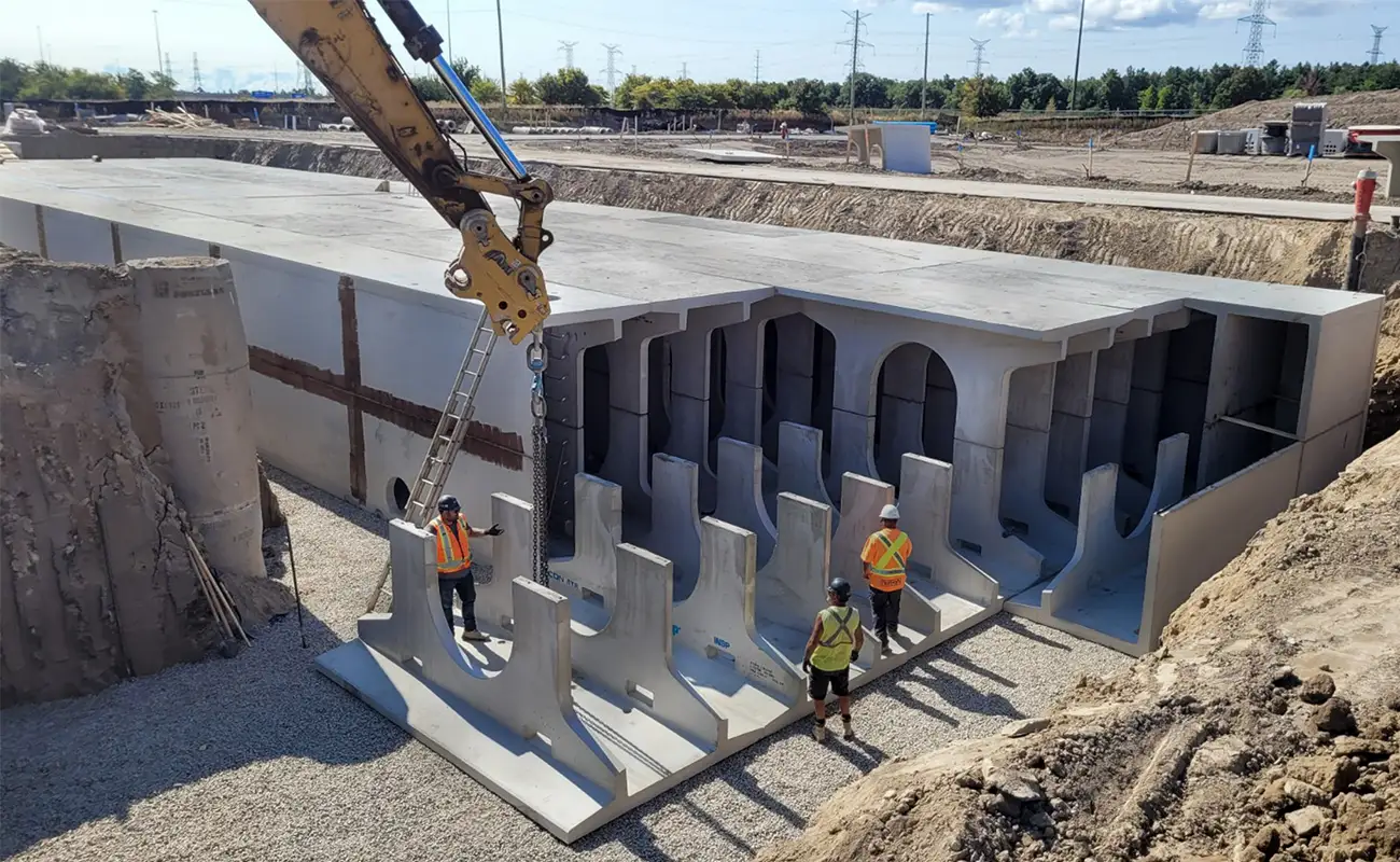Construction workers guide a large precast concrete structure being lifted by a crane at an excavation site with other sections nearby. Crane lowering StormTrap DoubleTrap precast concrete unit into excavation at 5150 Ninth Line Mississauga Ontario