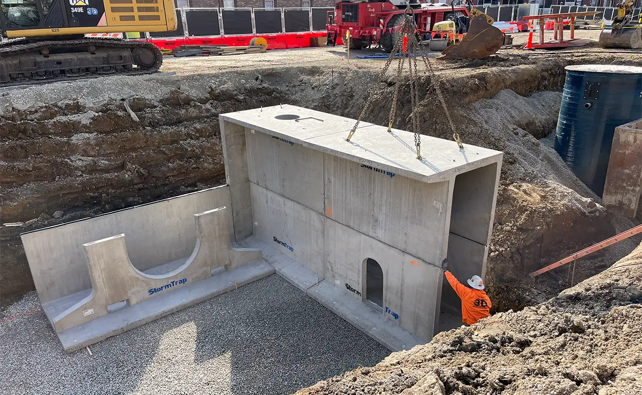 A worker guides a large precast concrete structure being lowered into an excavated construction site by a crane. Construction equipment and barriers are visible in the background.