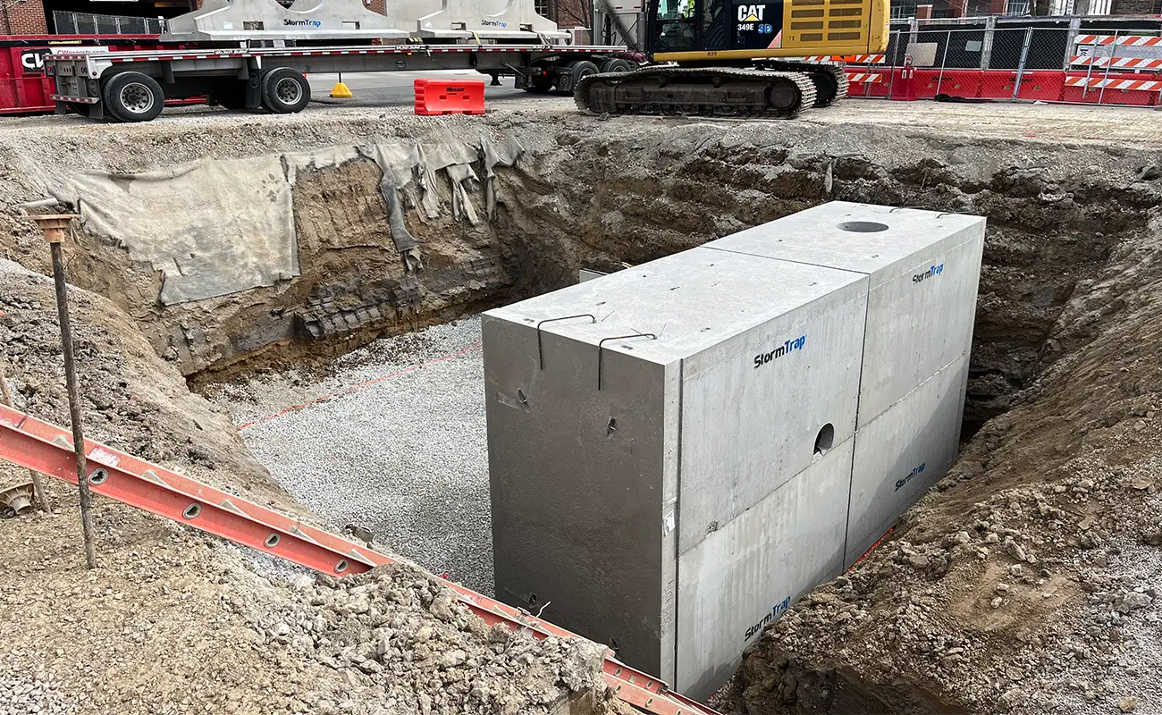A large rectangular concrete structure is installed in an excavated pit at a construction site, with gravel spread at the pit’s base and heavy machinery in the background.