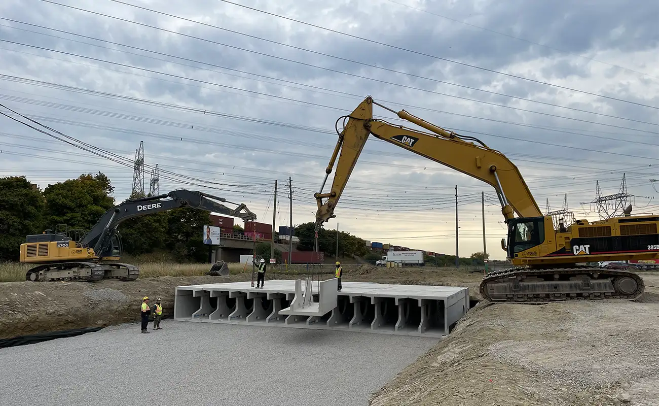 Wide shot of the excavation site. Multiple installed DoubleTrap units are visible in a row. Two workers stand at the edge of the system. A CAT excavator are both on site. Transmission towers and construction visible in background.
