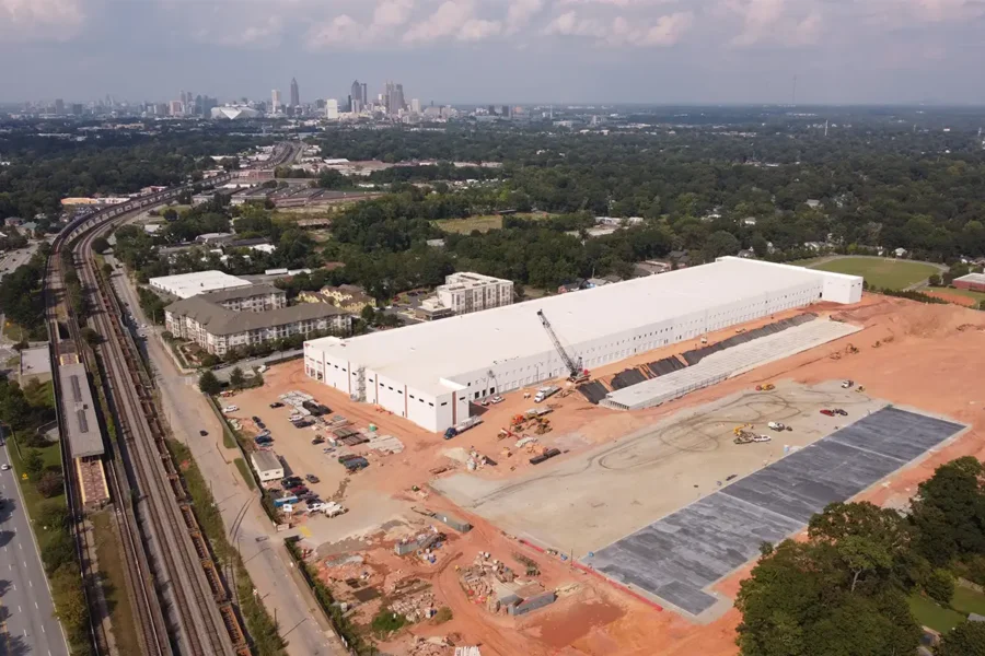 Aerial view of a large warehouse under construction with parking areas being developed, surrounded by trees and city buildings visible in the distant background.