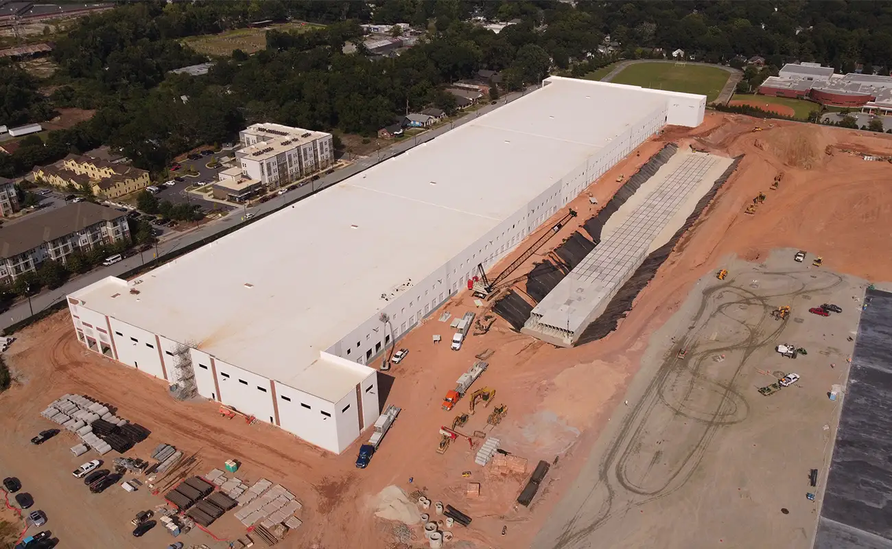 Aerial view of a large white industrial warehouse under construction, surrounded by dirt, construction materials, and vehicles. Residential buildings and trees are visible in the background.