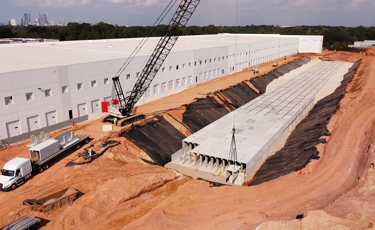 A construction site with a crane placing large concrete panels in a trench beside a white industrial building, with trucks and equipment nearby.