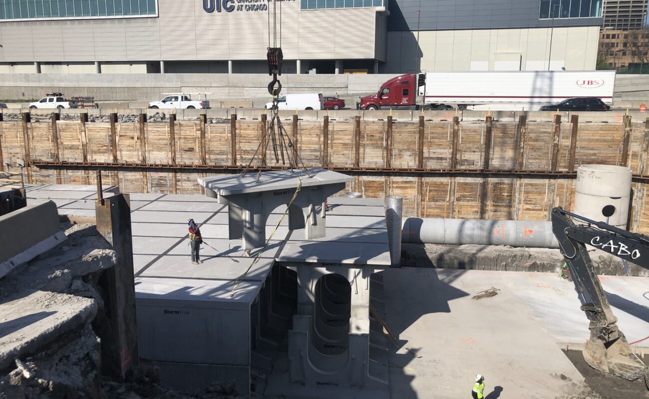 Construction site with workers, crane lifting a large concrete structure, and an excavator near a deep foundation pit; UIC building visible in background.