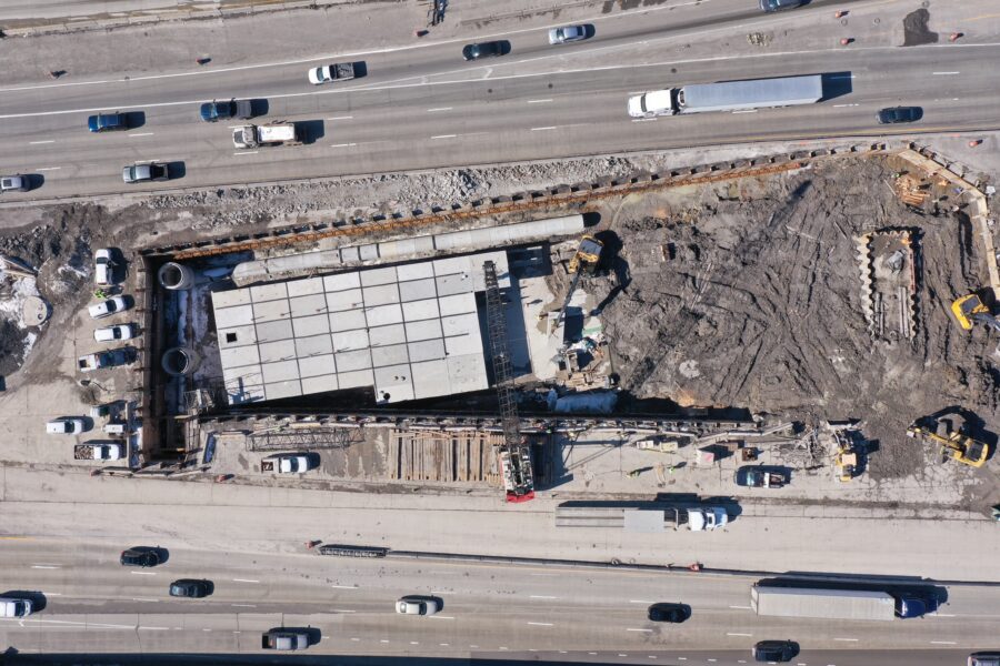 Aerial view of a construction site between two busy highways, with vehicles passing and construction equipment working on the site.