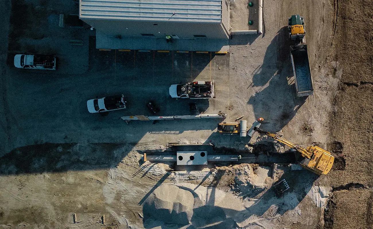 Aerial view of construction site with excavator digging, trucks parked nearby, and workers operating machinery next to a building.