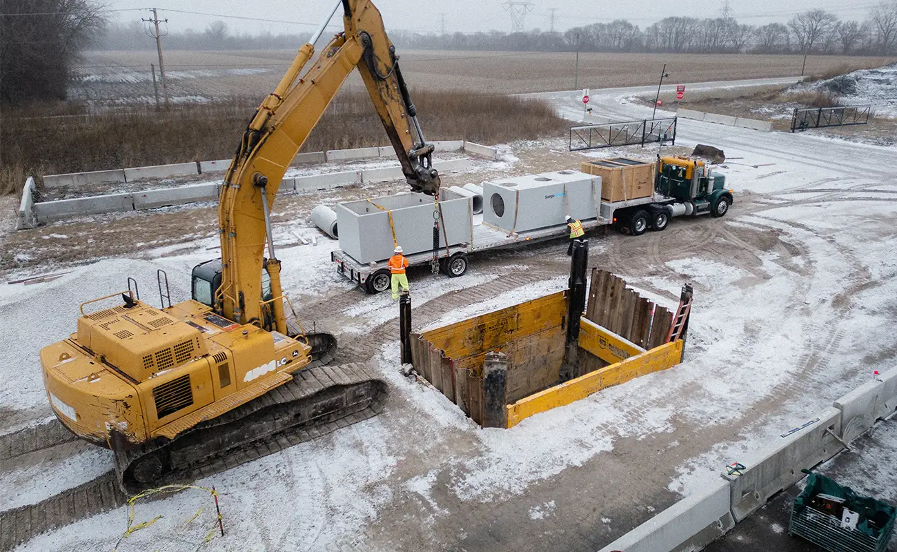 Construction workers guide a large yellow excavator lifting a concrete structure near an open trench at a snowy worksite with a truck nearby.