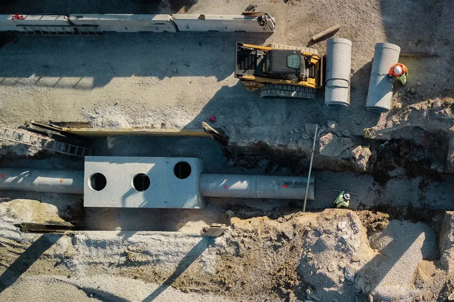 Aerial view of a construction site with workers installing large concrete pipes and rectangular structures using heavy machinery.