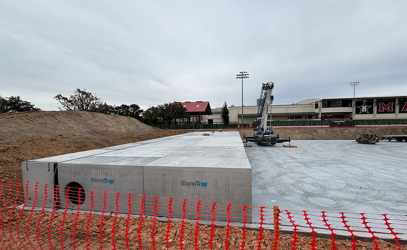 A construction site with large StormTrap concrete units installed, surrounded by dirt piles and orange safety fencing; buildings and light poles are visible in the background.