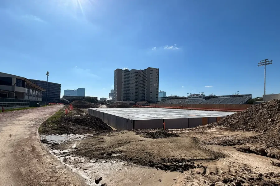A construction site with a concrete foundation in progress, surrounded by dirt piles, near a stadium and tall buildings under a clear blue sky.