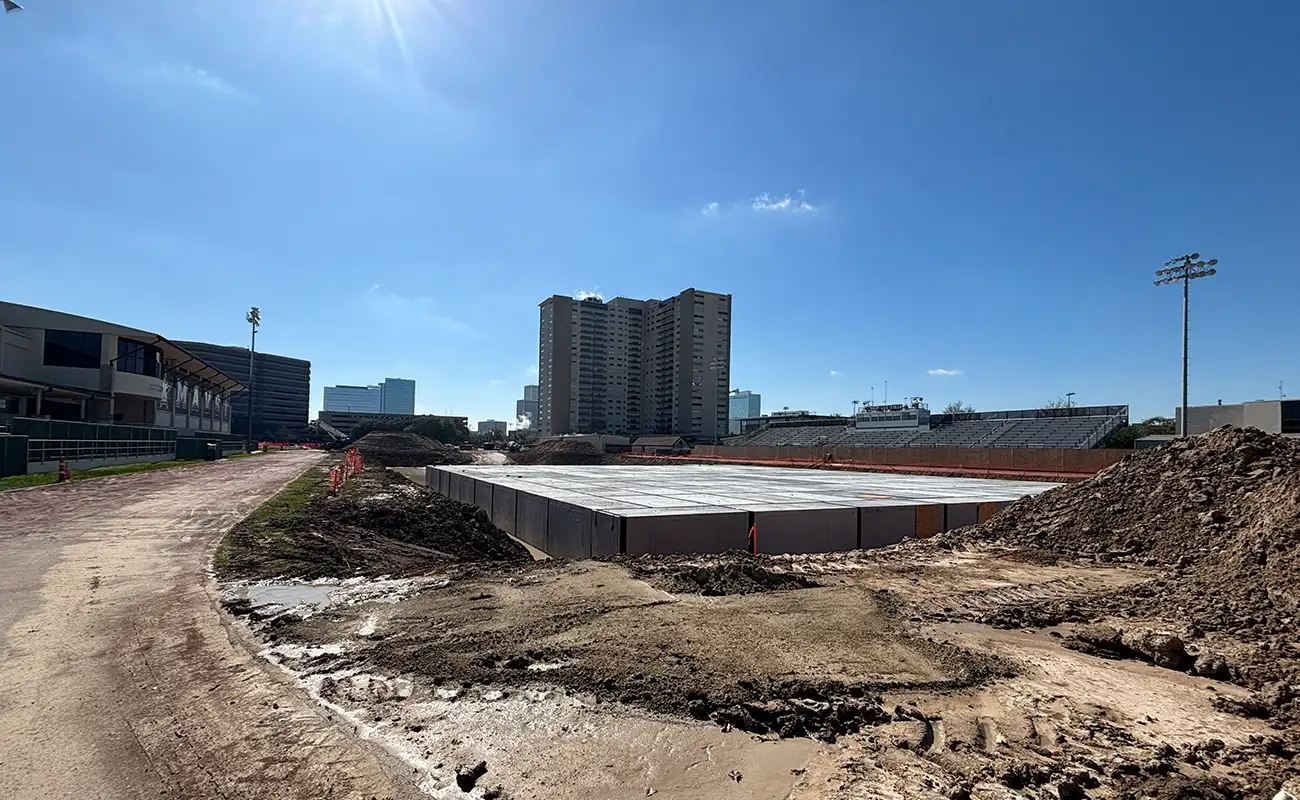 A construction site with a concrete foundation in progress, surrounded by dirt piles, near a stadium and tall buildings under a clear blue sky.