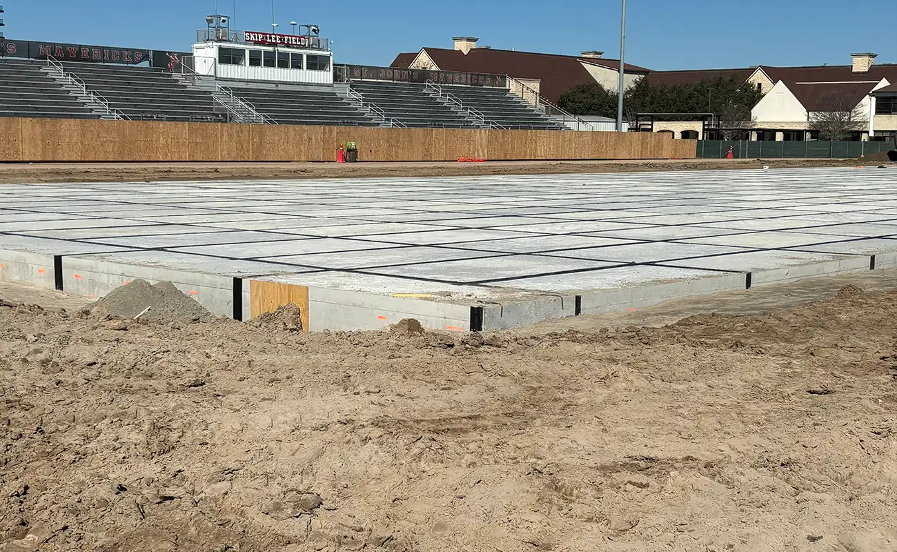 A sports field under construction with a grid of concrete slabs in the foreground, bleachers and buildings visible in the background.