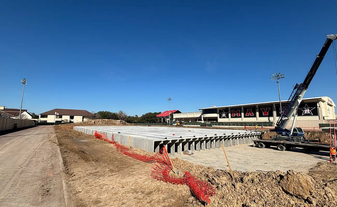 Construction site with concrete beams and a crane near a stadium and buildings under a clear blue sky. Orange safety netting lines the dirt area.