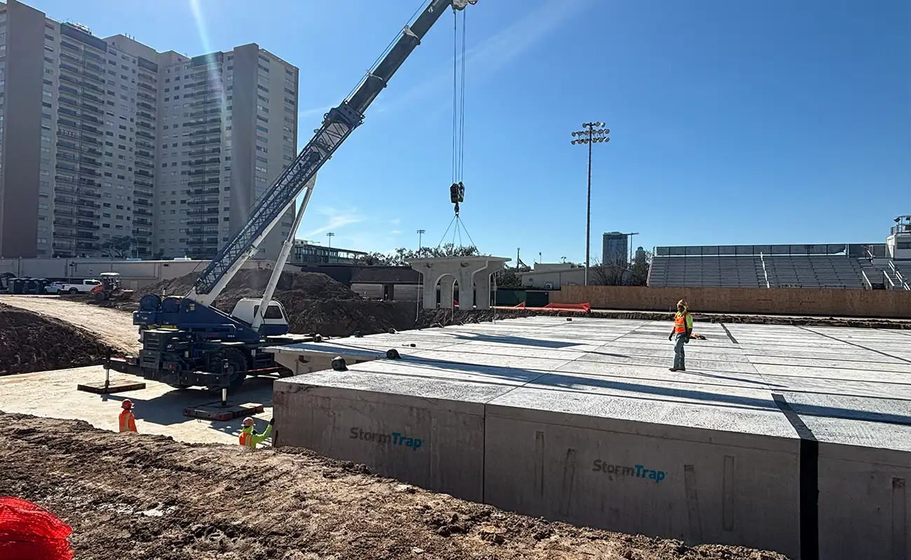 A crane lifts a large concrete slab at a construction site with workers nearby; large buildings and a stadium are visible in the background.