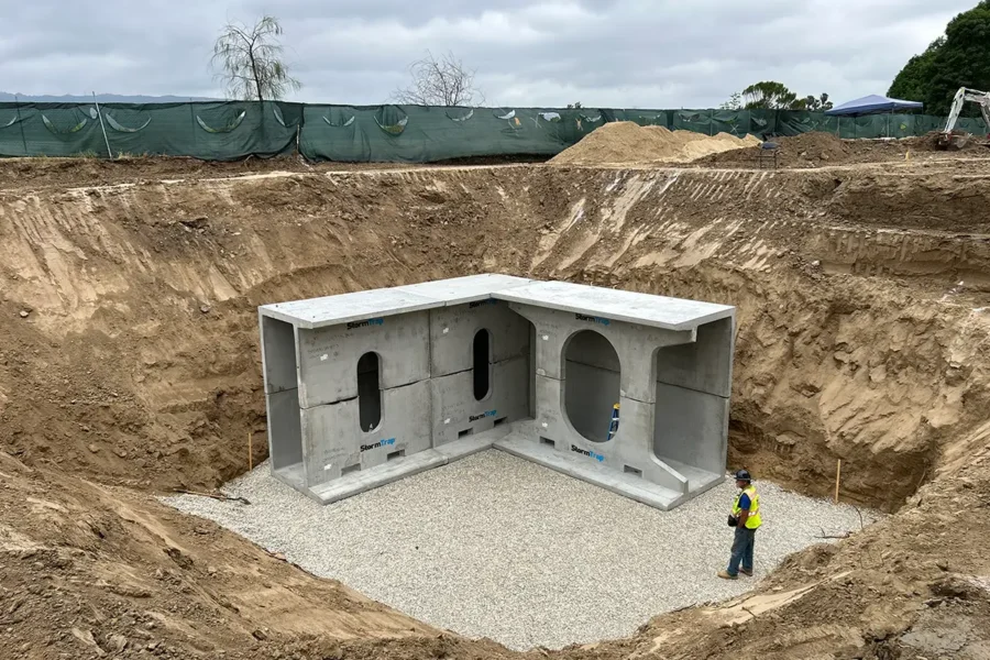A construction worker stands in a large excavated pit with partially installed concrete stormwater infrastructure and gravel base at a construction site.