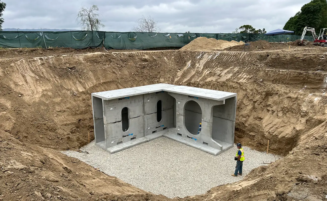 A construction worker stands in a large excavated pit with partially installed concrete stormwater infrastructure and gravel base at a construction site.