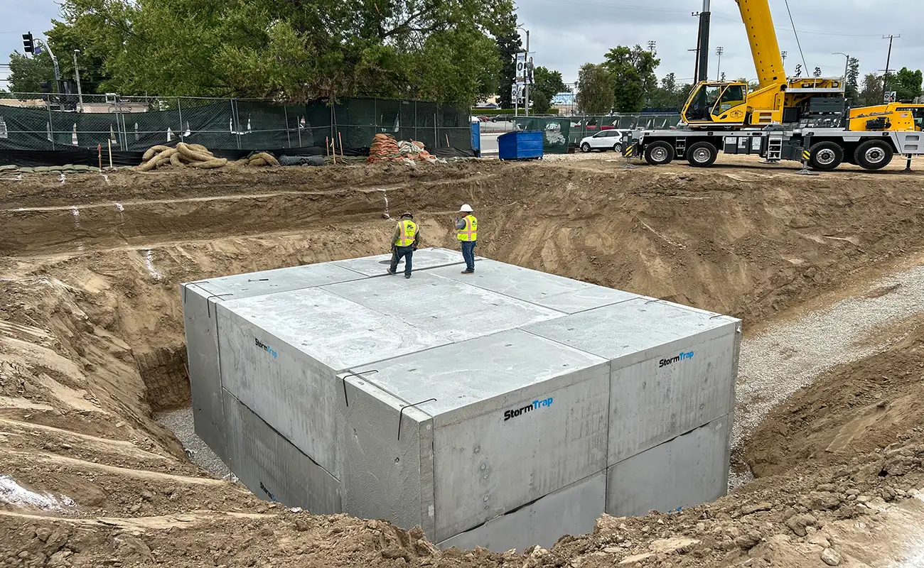 Two construction workers stand on top of large concrete blocks in an excavated site, with a crane and fencing visible in the background.