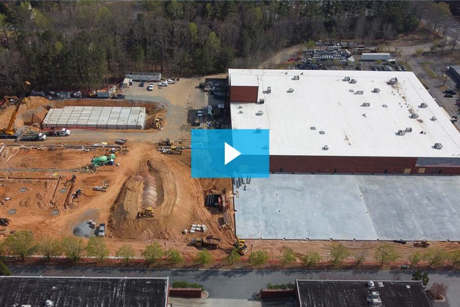 Aerial view of a construction site showing a large white-roofed building, adjacent cleared land with construction equipment, and surrounding trees.
