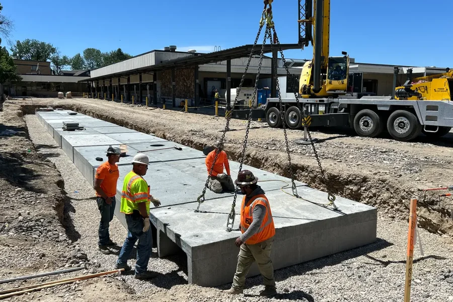 Four construction workers guide large concrete slabs into place with a crane at an outdoor construction site on a clear day.