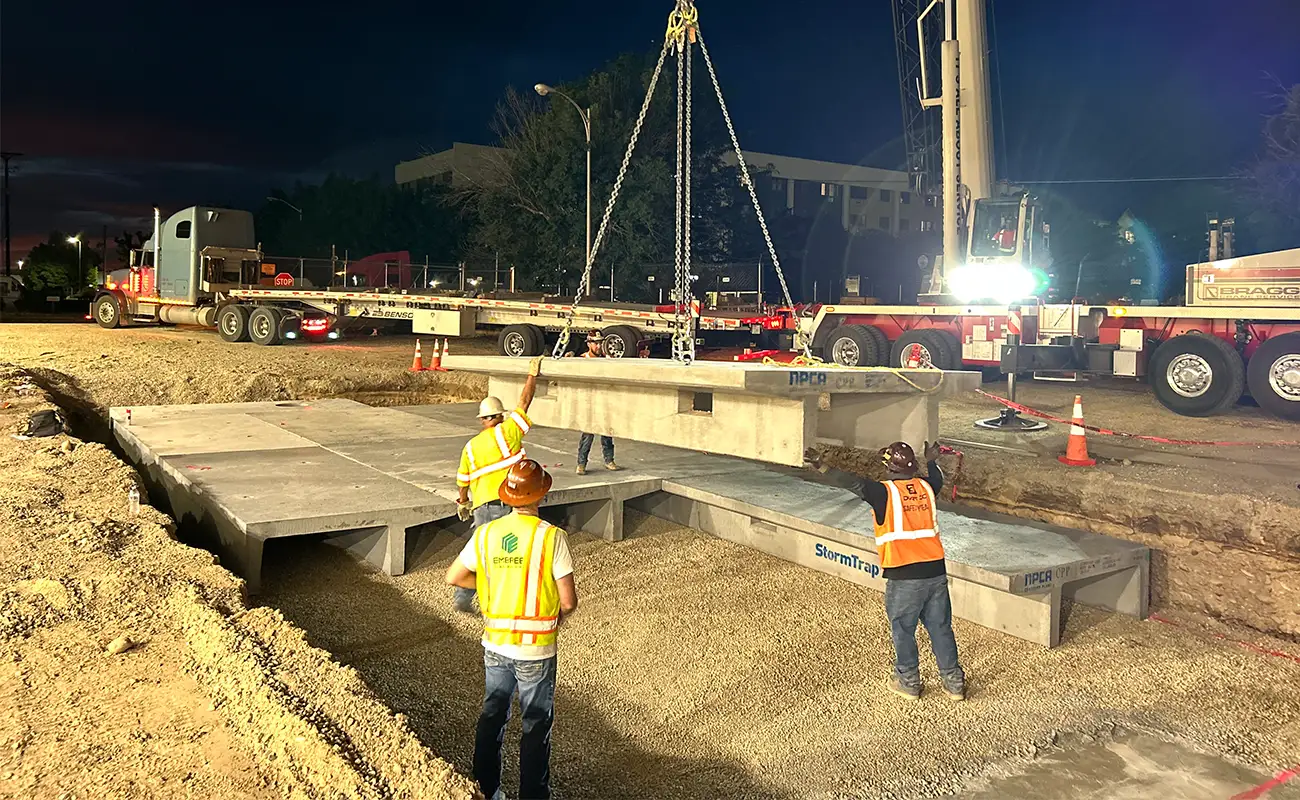 Construction workers guide a large concrete slab being lowered by a crane into position at a worksite at night.