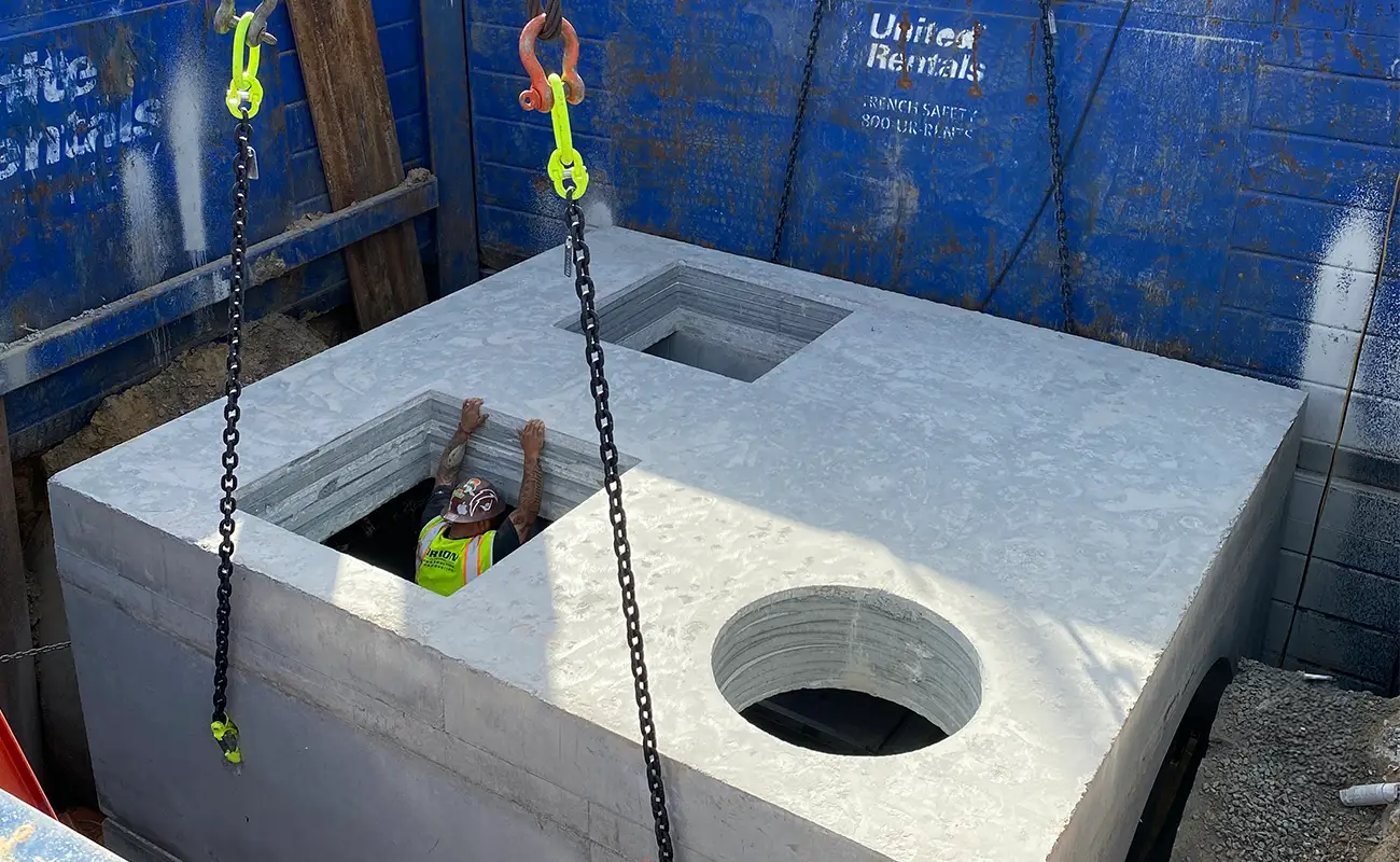 A construction worker stands inside a large concrete box structure, raising their hands through a square opening, with lifting chains attached to the box.