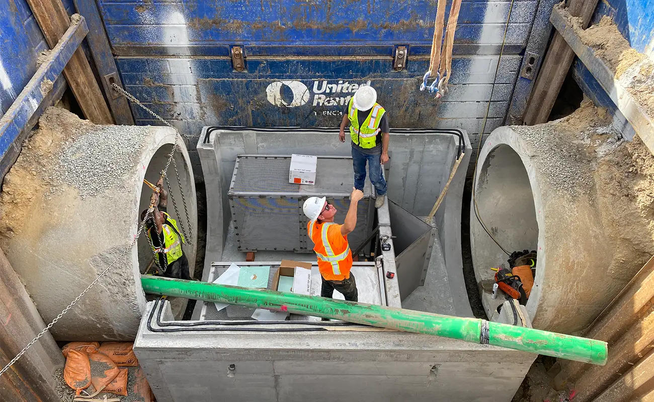 Three construction workers in safety gear stand inside a concrete utility vault between two large pipes at an active construction site.
