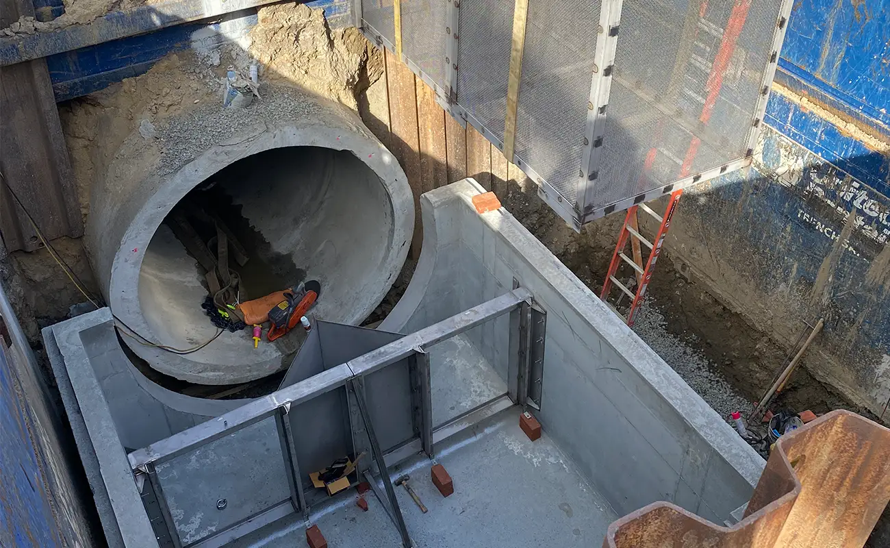 A worker in safety gear inspects a large concrete pipe section at a construction site with metal supports, a ladder, and dirt excavation visible.