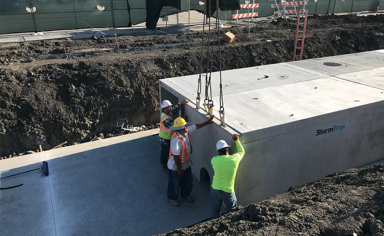 Three construction workers guide a large precast concrete StormTrap module being lowered by a crane into a detention system trench in Frisco, Texas.