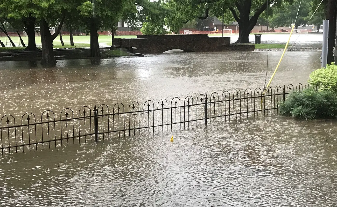 A flooded park before an underground stormwater detention system was installed, featuring a submerged fence and a stone footbridge in the background during heavy rain.