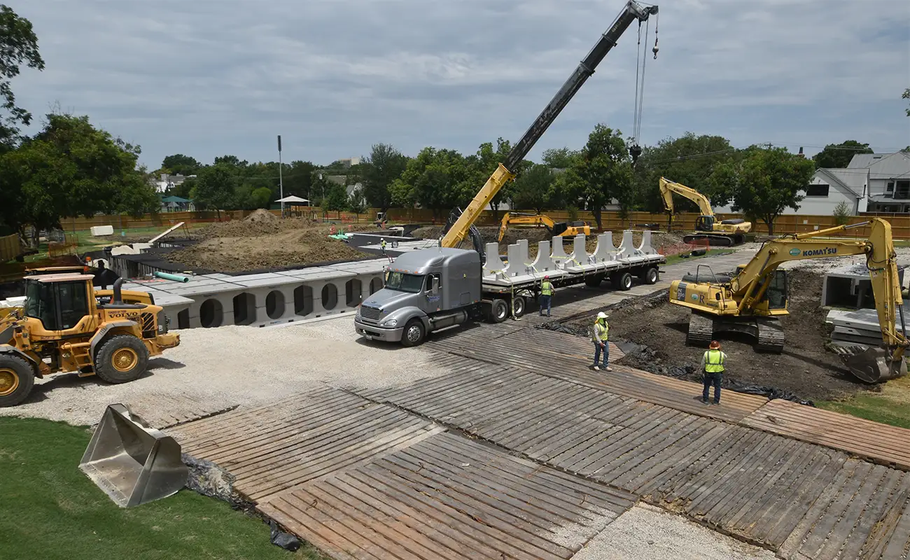 Construction site for an underground stormwater detention system in University Park featuring a mobile crane, a flatbed truck hauling concrete barriers, two excavators, and workers on ground mats.